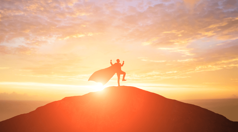 Un homme au sommet d'une montagne face à un coucher de soleil avec une cape de super-héros. Ayez confiance en vous pour apprendre l'anglais et dîtes “Now I Speak” !