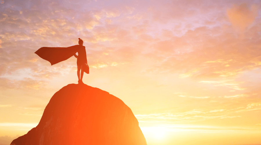 Une femme d'affaire au sommet d'une colline devant un coucher de soleil avec une cape de super-héroïne. Ayez confiance en vous pour apprendre l'anglais et dîtes “Now I Speak” !